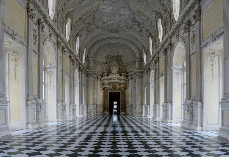 The main ballroom of an Italian castle surrounded in hand molded white walls with a checkerboard floor.
