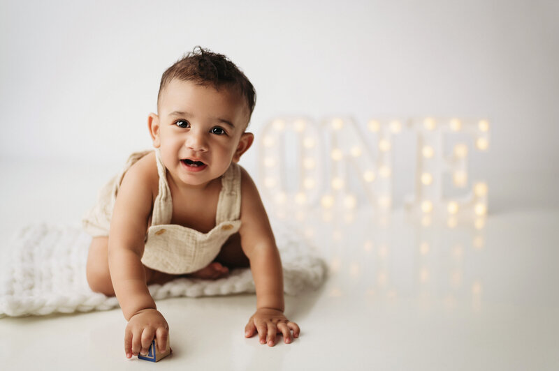 one year old baby boy wearing neutral overalls and crawling towards the camera and smiling in front of white backdrop and lit up ONE sign photographed by Milford Cake Smash Photographer Drea S Photo