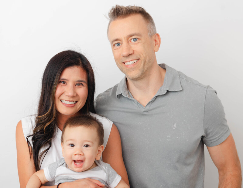 Family photo with mom and dad against a white backdrop with mom holding six-month-old baby. 