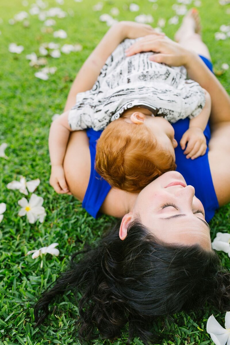 A mother holds her son against her chest, laying on the ground with flowers all around.