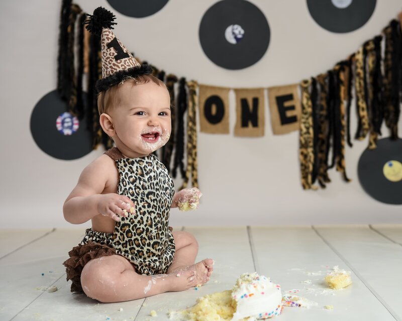 A smiling baby girl in a leopard print outfit and birthday hat, sitting in a rock-and-roll-themed cake smash setup with vinyl records and a "ONE" banner in the background.
