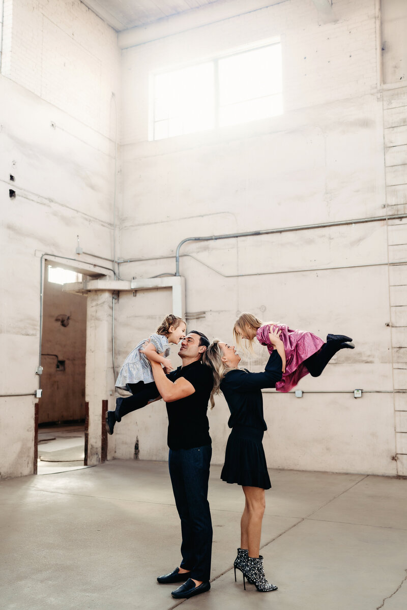 Stylish family with two young daughters swings them around in a trendy warehouse during winter photo session in downtown Denver, Colorado.