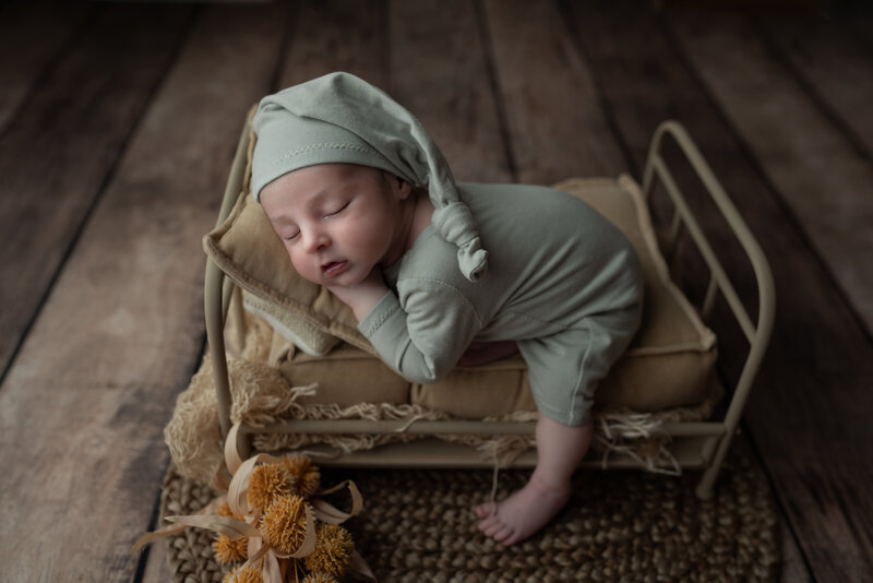 A softly sleeping newborn dressed in sage knitwear on a mini rustic bed styled with vintage textures.