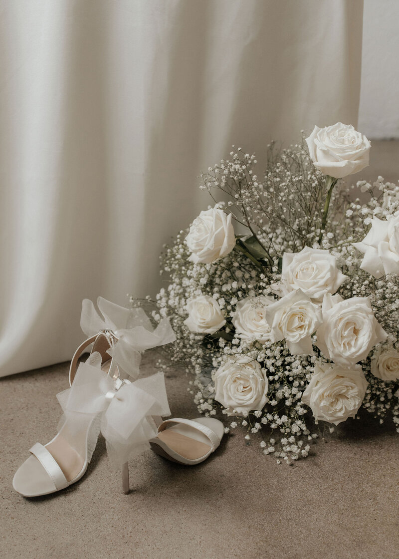 white heels with bows on them sitting in front of a white rose and baby's breath bouquet