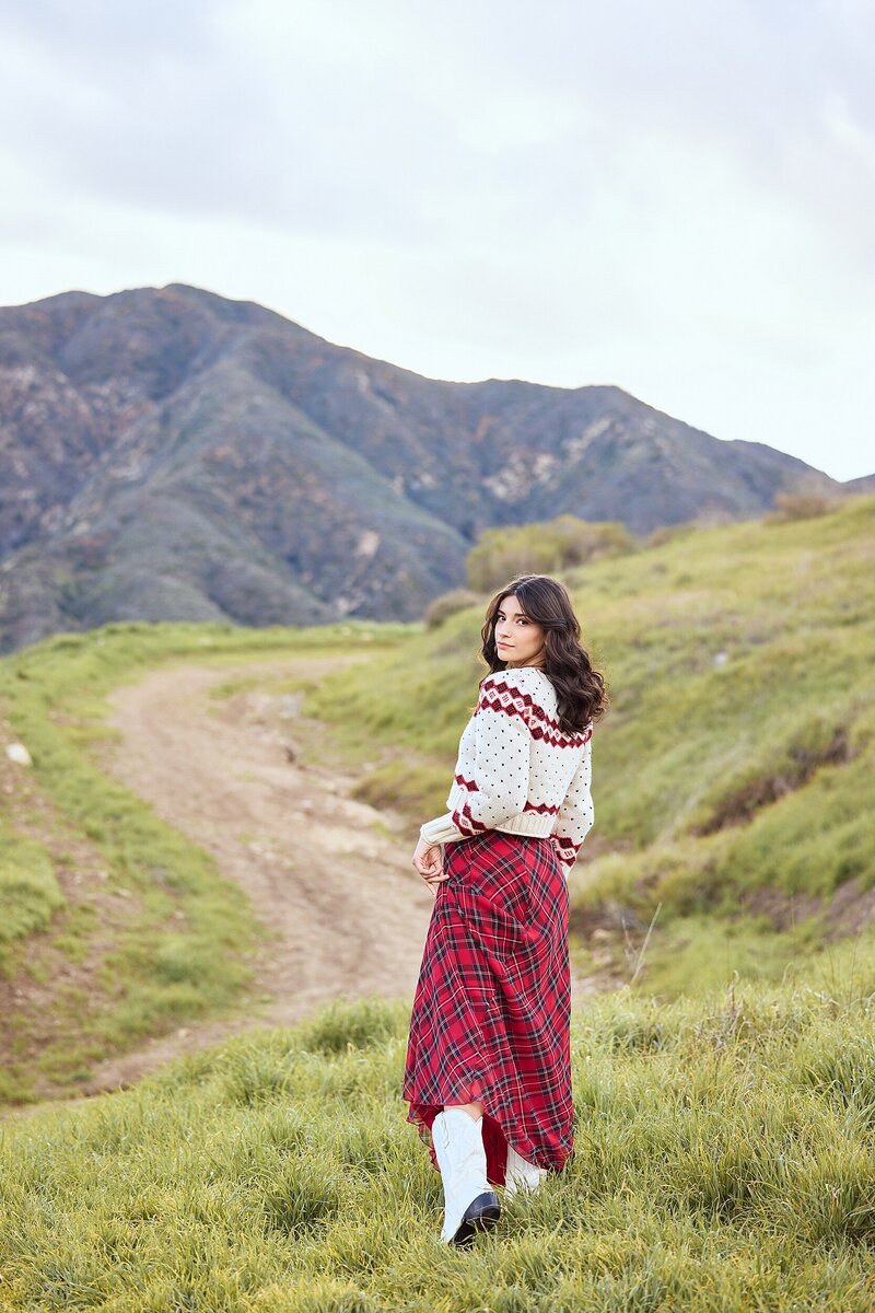 Senior girl walking through a scenic trail in the mountains, wearing a red plaid skirt and white boots with a cozy sweater—perfect blend of editorial and outdoorsy.