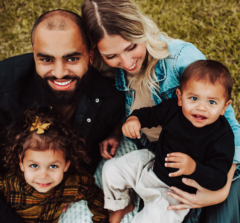 mom is looking at little boy, dad and little girl is looking at the camera, family is snuggling, happy family, sitting on the grass