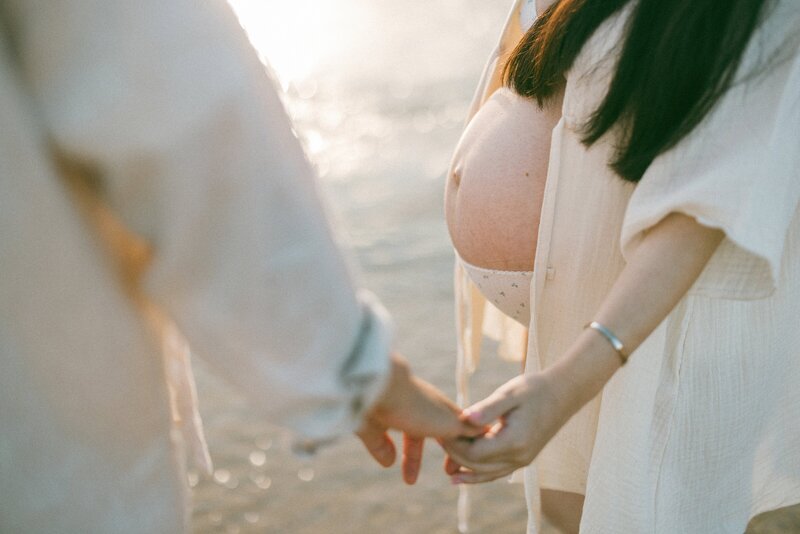 A light filled image of a couple holding hands, captured during a maternity session at the beach with Tealily