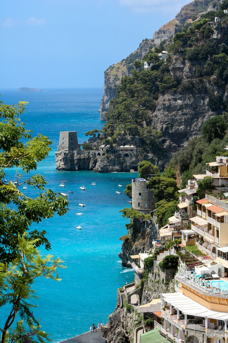 Bright midday view of the Amalfi Coast with turquoise waters, historic stone watchtowers, and pastel hillside villas overlooking boats scattered across the bay.