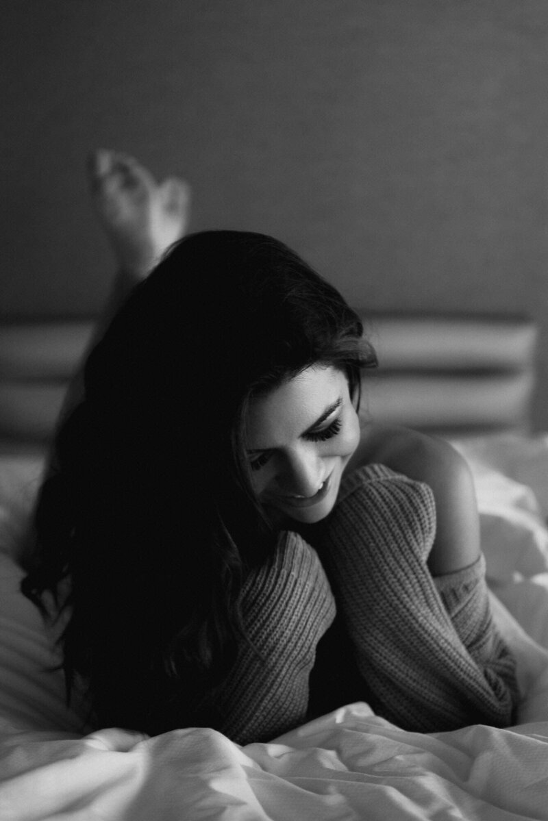  Phoenix boudoir photography of a woman laying on a bed in a cozy sweater, smiling softly in a black and white portrait that captures warmth and confidence.
