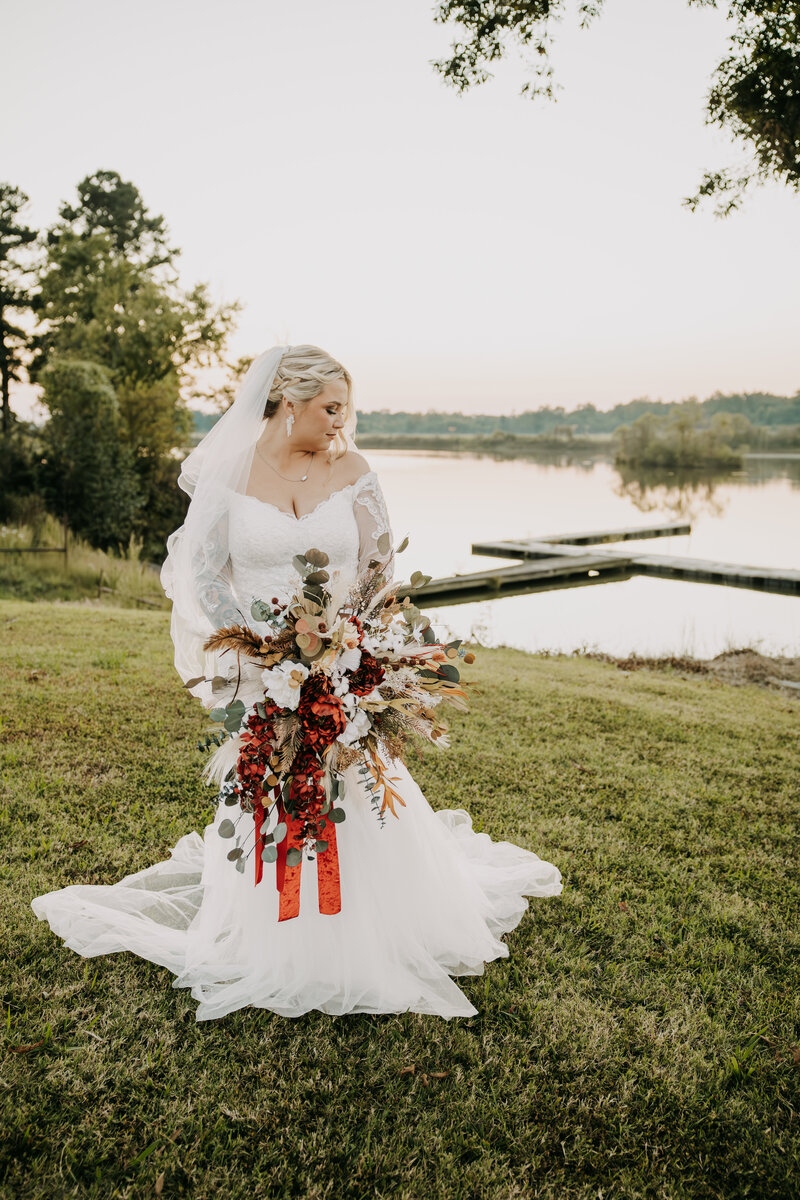Bride looks down her shoulder at her bridal bouquet, standing in front of a lake with a dock at sunset.