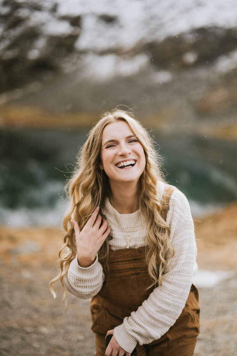 alaska elopement photographer with long blong hair and carhartt overalls standing in front of a glassy lake in hatcher pass