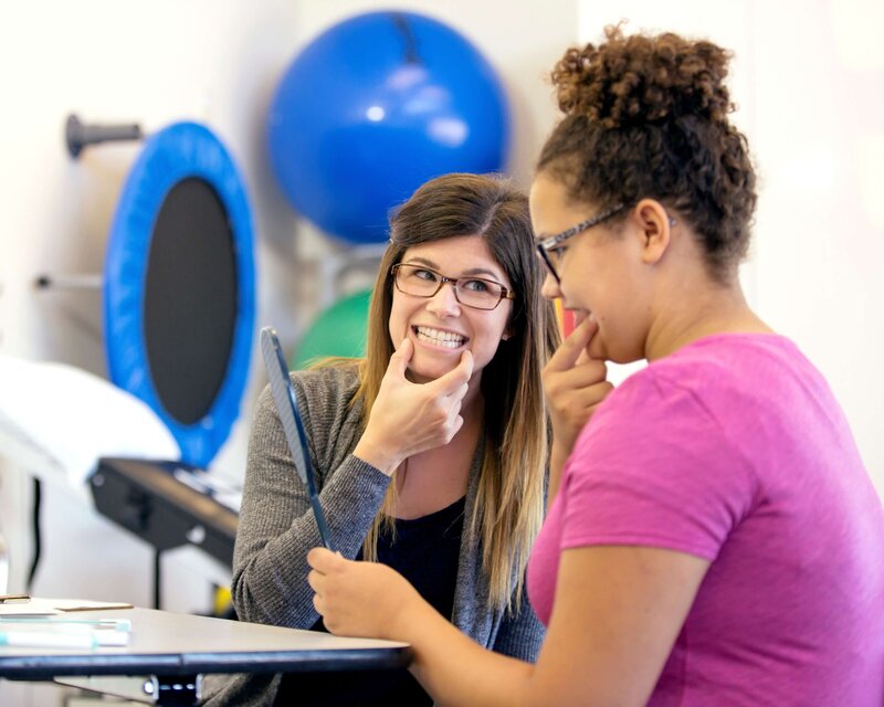 Speech-language pathologist guiding a teenage girl through articulation exercises using a mirror during a speech therapy session.