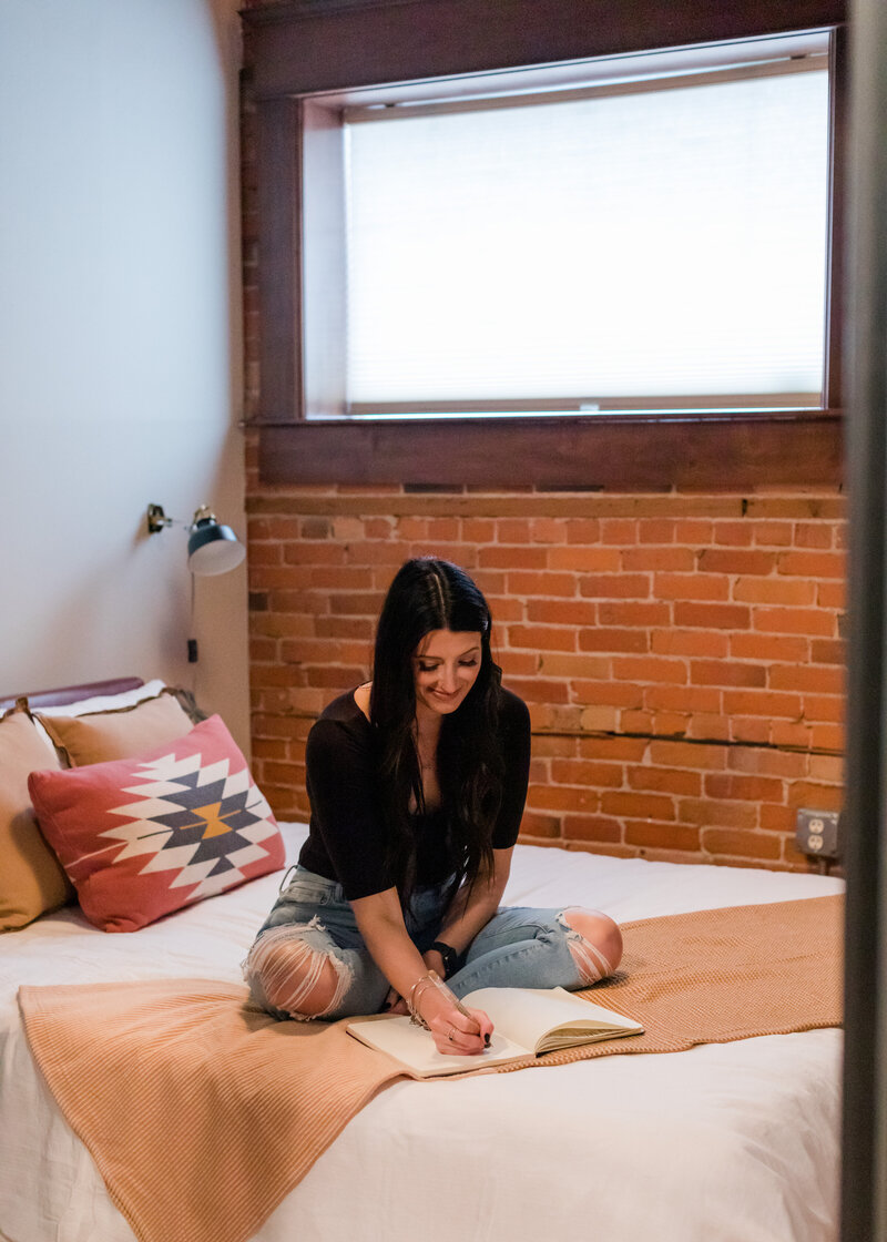 Allie Clark writing in a notebook while sitting on a bed, representing the brand discovery phase for Allie Clark Designs.