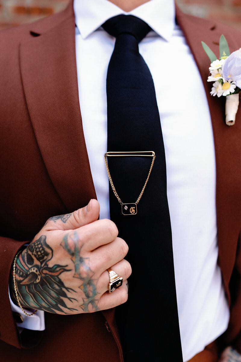 Close up detail photo of  groom's maroon suit. His tattood hand is holding his lapel and he had a G pin on his black tie.