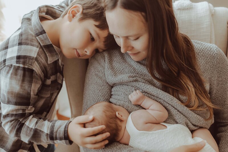 A mother holds a baby while a young boy gently touches the baby's head. The scene is warm and intimate, with the family seated comfortably at home.