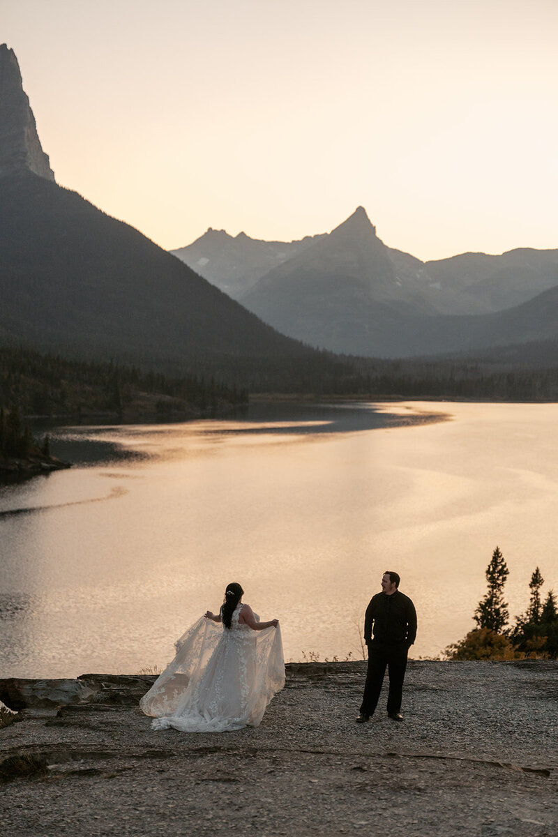 A bride and groom stand together overlooking a golden lake at sunset surrounded by mountain peaks in Glacier National Park, captured by Sydney Breann Photography during their romantic Montana elopement.