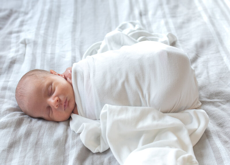 A peaceful sleeping newborn baby swaddled in soft white fabric rests on light bedding during a lifestyle newborn session in Bergen County, NJ