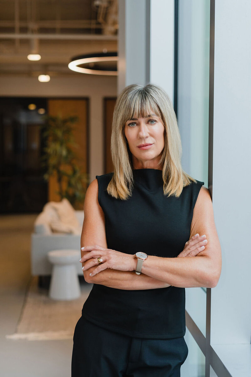 Female Brightdot employee in black tank top with shoulder length blonde hair stands with arms crossed inside modern corporate office setting | Sara Coffin Photo in NC