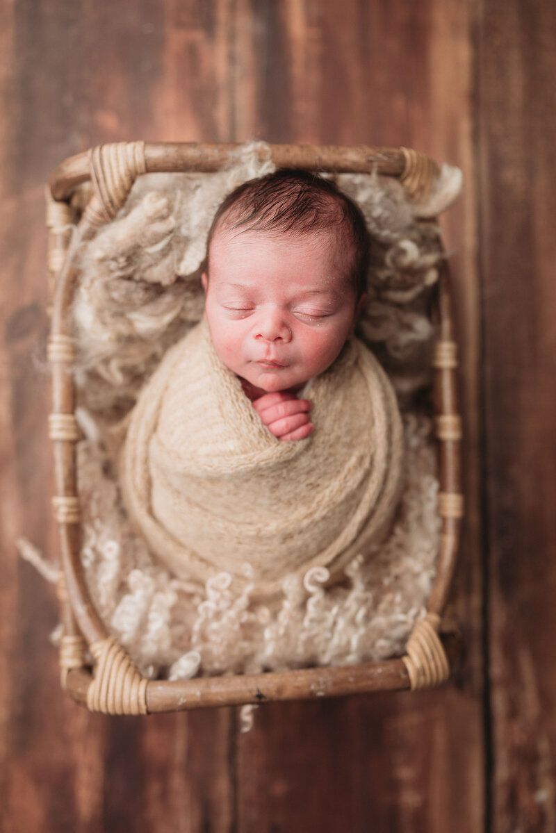 Newborn boy wrapped in taupe knit swaddle laying on faux fur in a basket on brown wood floor