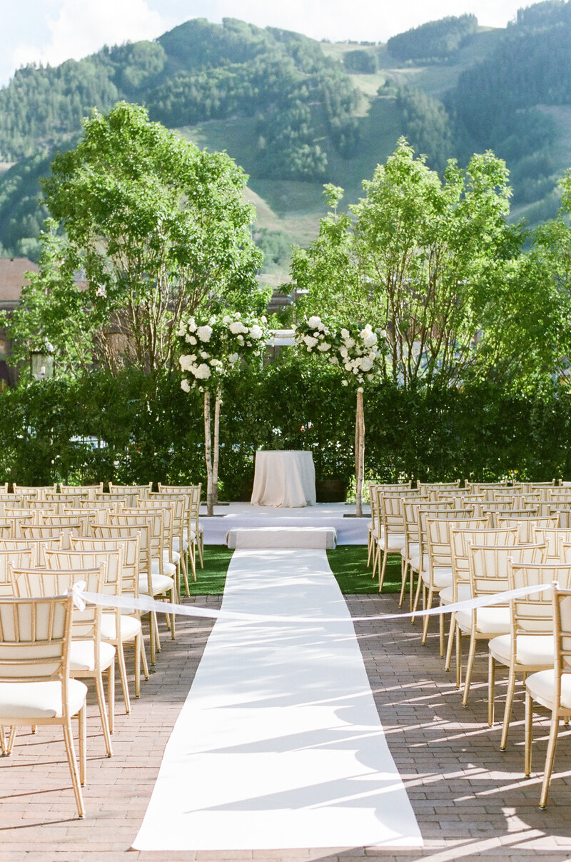 Empty outdoor wedding ceremony space, lined with wooden chairs. There is a white aisle, with a floral chuppah at the end, there are mountains in the background.