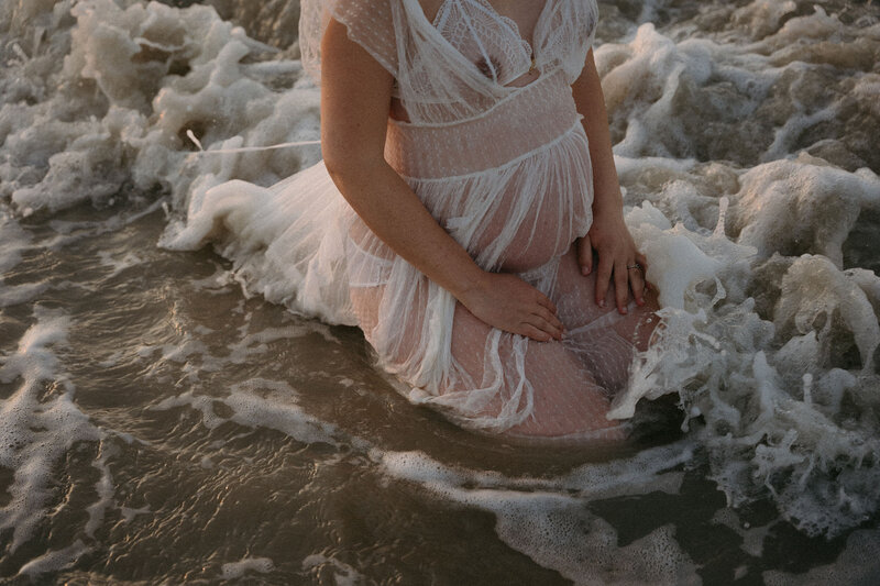 Femme au ventre arrondie et robe transparente agenouillée dans l'écume à la mer. Séance photo grossesse par Kamescop.