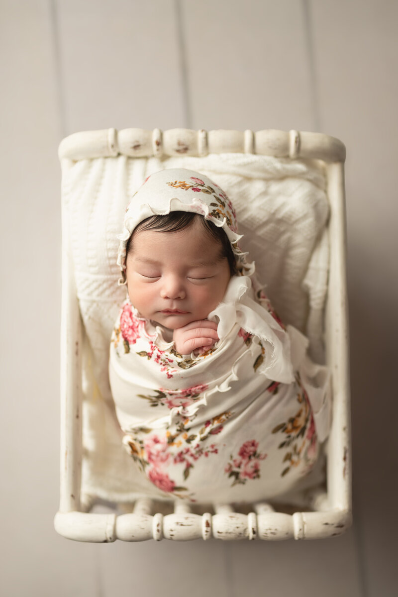 Newborn girl styled in full floral wrap and bonnet, posed on a soft cream bed in vintage tones.