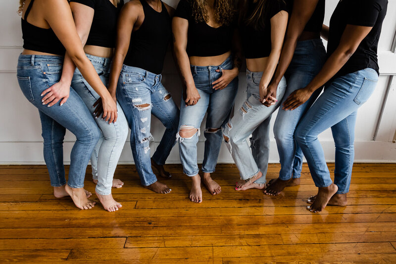 Seven girls of all skin tones wearing a black shirt and light blue jeans standing against a white wall holding hands