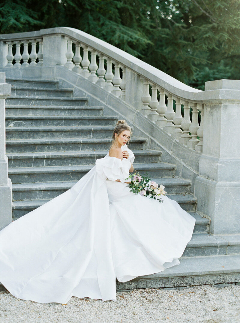 Dramatic bride in lace gown with long train wearing a pearl crown standing in front of carved wood church doors