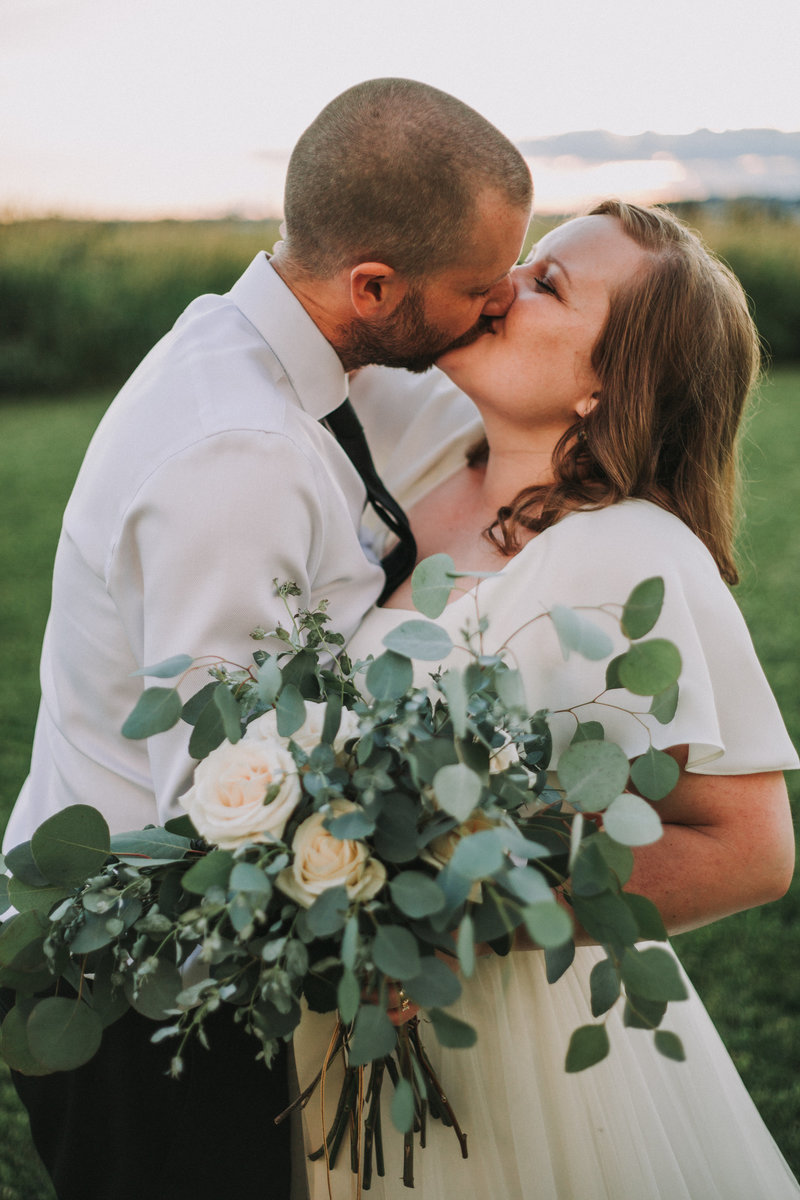 Laura-Anne and her husband kissing on their wedding day while she holds her bouquet of greenery