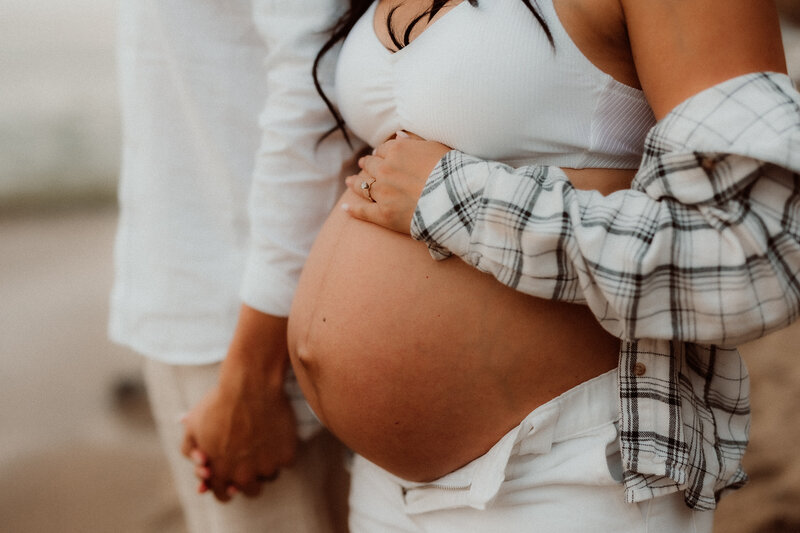 Close-up of a pregnant woman’s belly with her partner holding her hand, both dressed in neutral tones during an intimate maternity photoshoot in Southern California