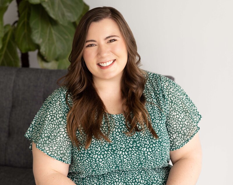 Smiling woman with long brown hair wearing a green patterned dress, sitting on a dark gray sofa in a bright studio space with a leafy plant behind her.