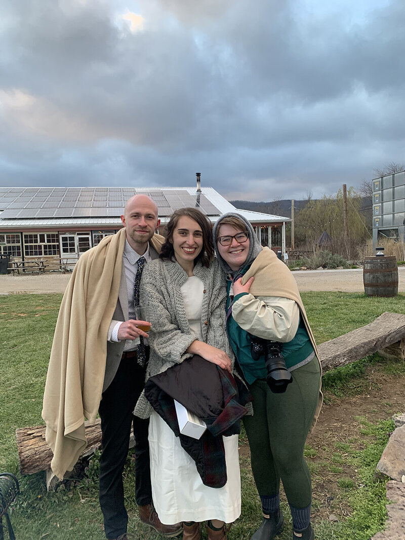 A Virginia elopement photographer stands next to an elopement couple outside of Rising Silo Brewery in Blacksburg, Virginia towards the end of their wedding day. The trio all wrap up underneath a blanket to keep warm during their chilly elopement day in March.
