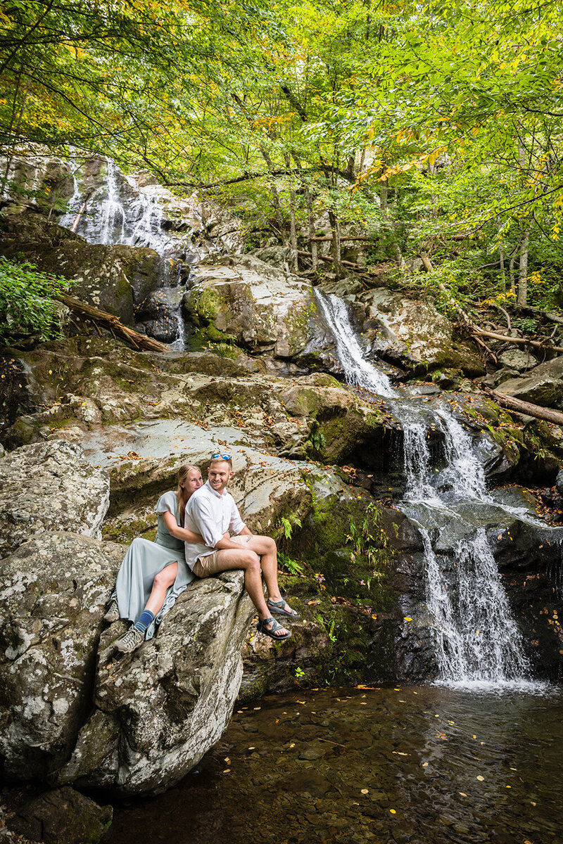 A couple on their elopement day sit on a large rock to the left of Dark Hallow Falls in Shenandoah National Park in Virginia.