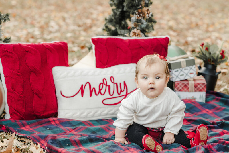 A baby dressed in a white sweater sits on a plaid blanket. Behind her are some Christmas pillows.