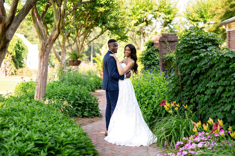 Bride and Groom in a garden embracing and smiling at the camera.  Garden florals in lilac, yellow and red surround them.