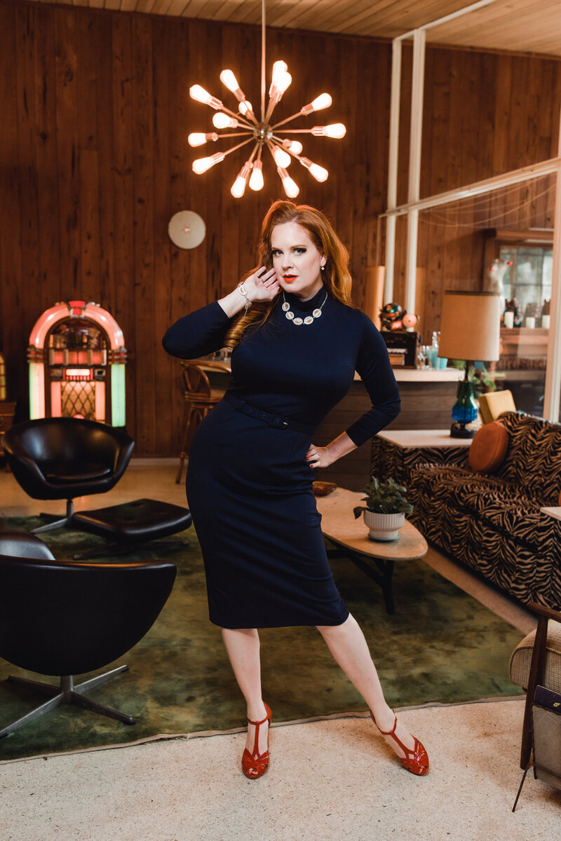 Classic pinup model in a form-fitting navy dress poses under a retro chandelier in a 1960s living room with bold vintage décor.