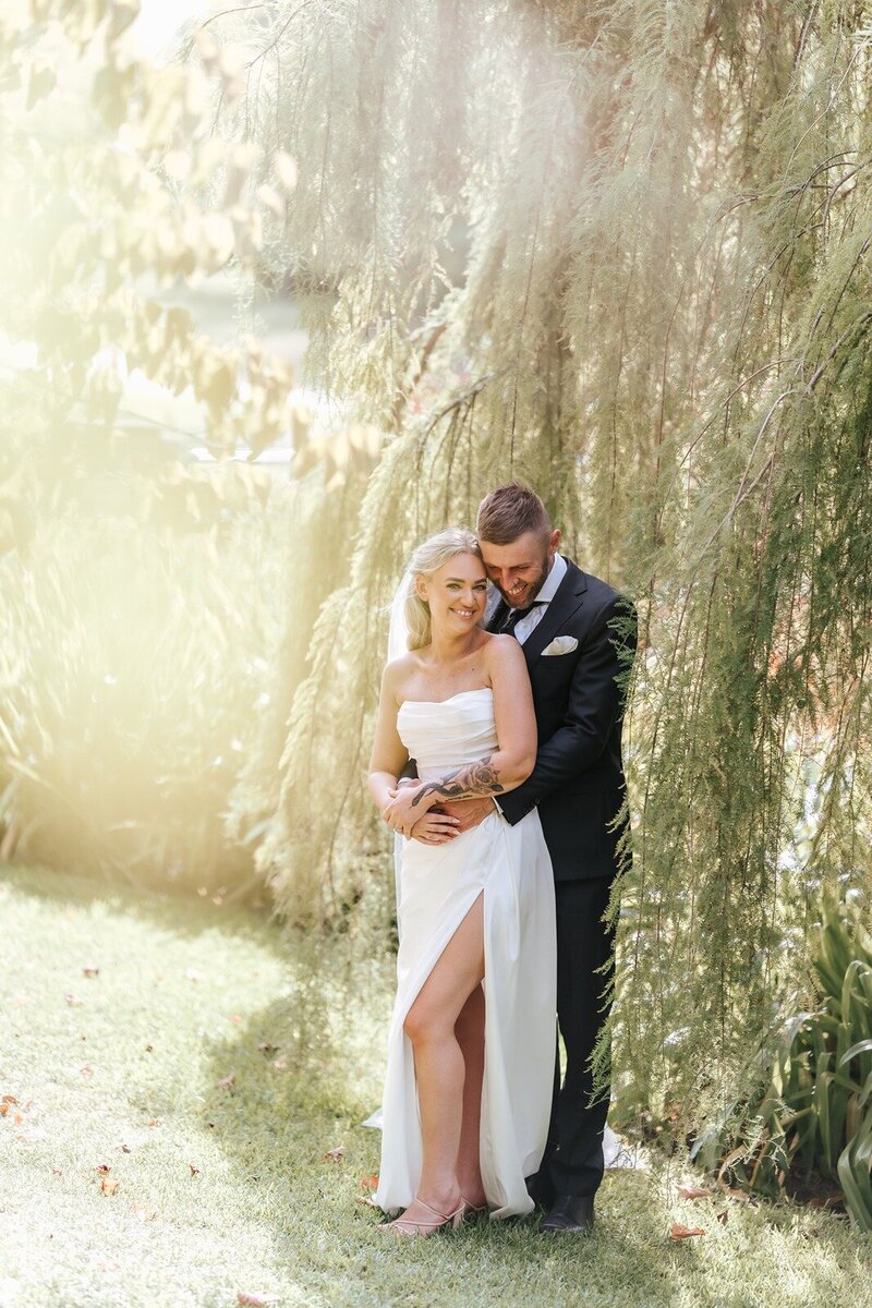 A bride standing in front of a tree with her groom holding her in golden light