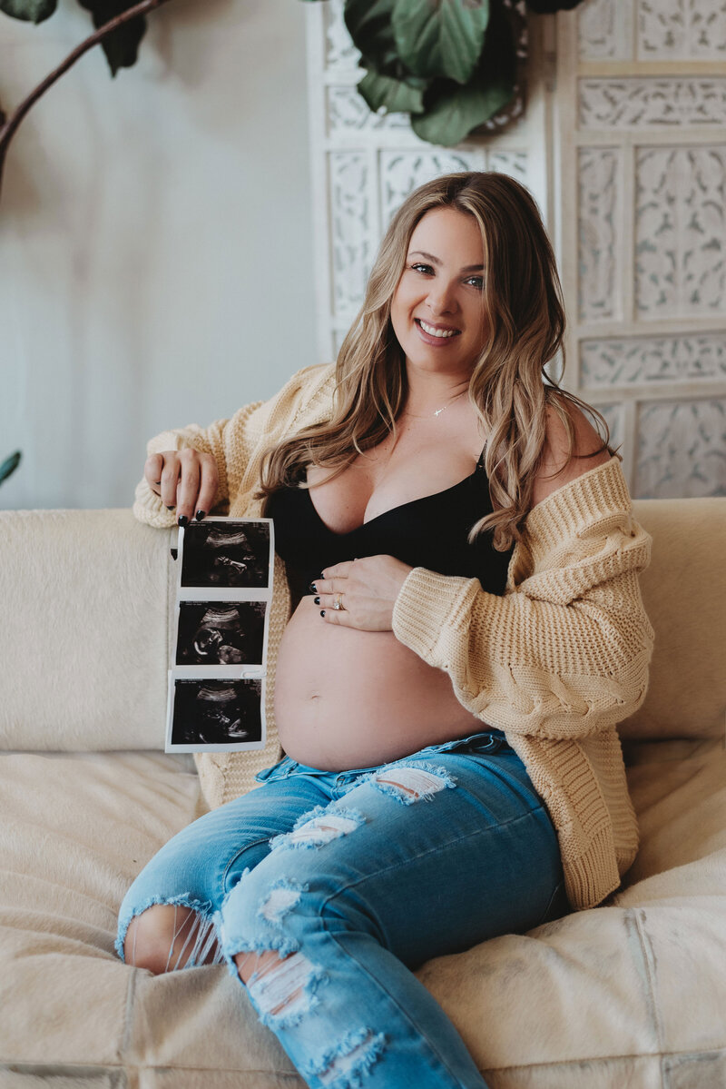 Pregnant woman in a crop top and sweater sitting on couch and holding ultrasound image during maternity photoshoot in Connecticut
