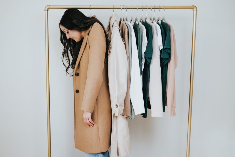 Woman with long, brown hair wears a camel-colored trenchcoat that hangs from a hanger on a clothing rack.