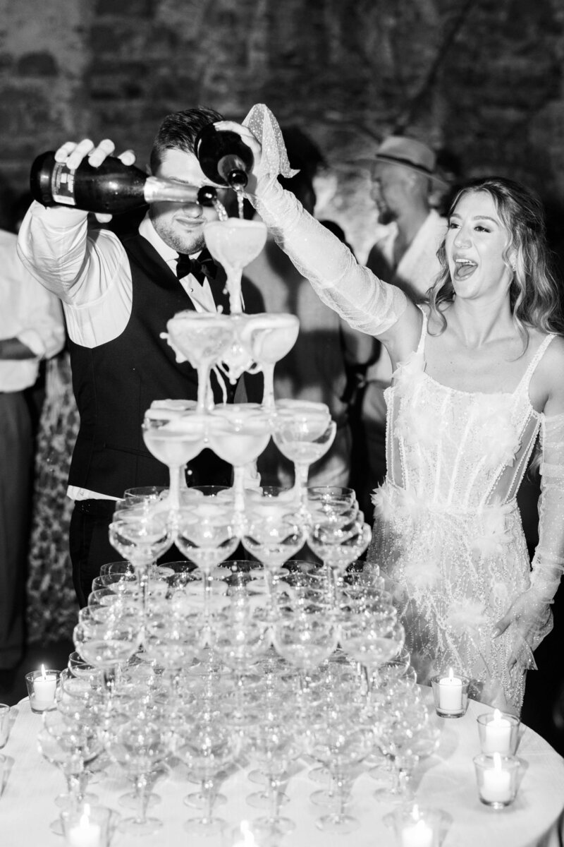 Black and white photo of bride and groom joyfully pouring champagne into a tower of glasses during their wedding reception.