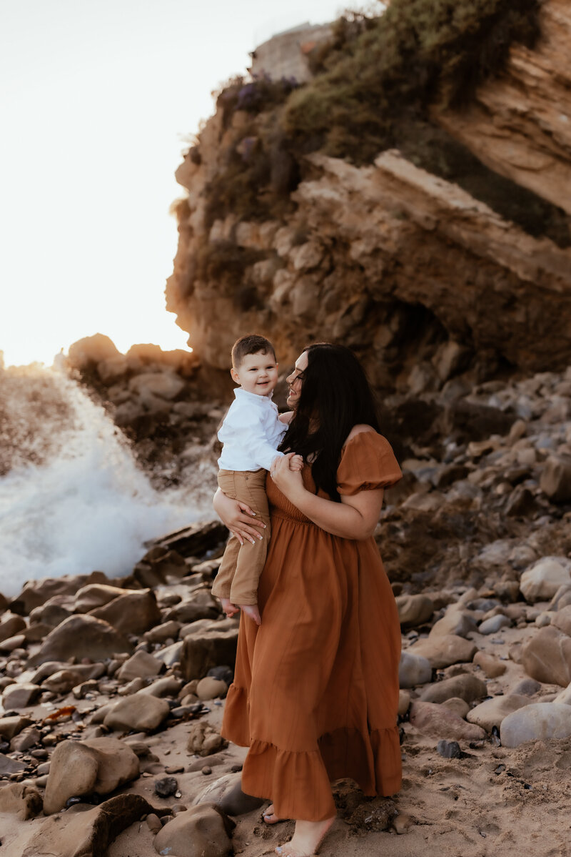 A mother wearing a long flowy rust colored dress holding her toddler boy as the waves crash on the rocky Newport Beach. 