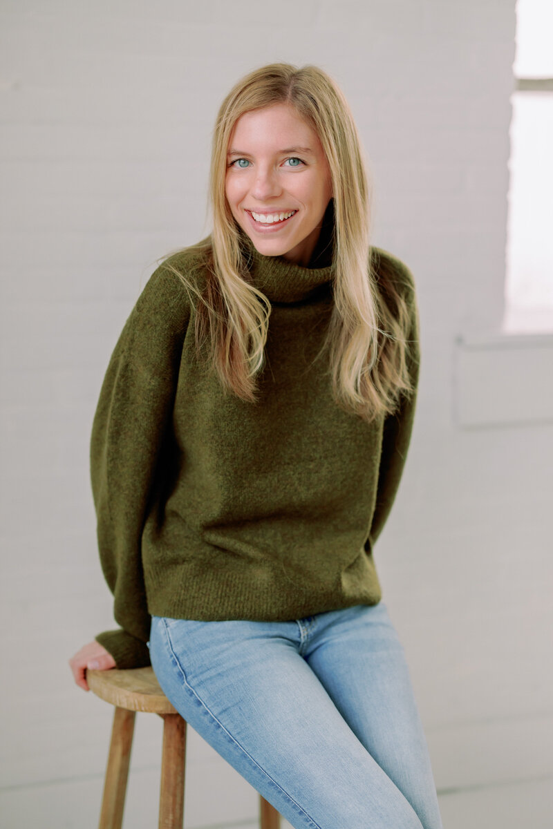 Nikki smiles while sitting on a stool, a white background behind her.