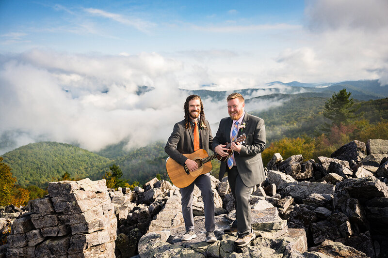 An LGBTQ+ couple stands on a pile of rocks at Blackrock Summit. Each of them hold a stringed instrument and smile towards their Virginia elopement photographer for a photo.  They are photographed at Blackrock Summit in Shenandoah National Park.