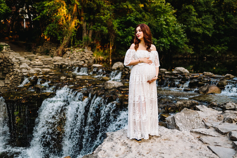 Woman in long white dress pregnant looking to the left side standing in a river. Behind are trees with rocks