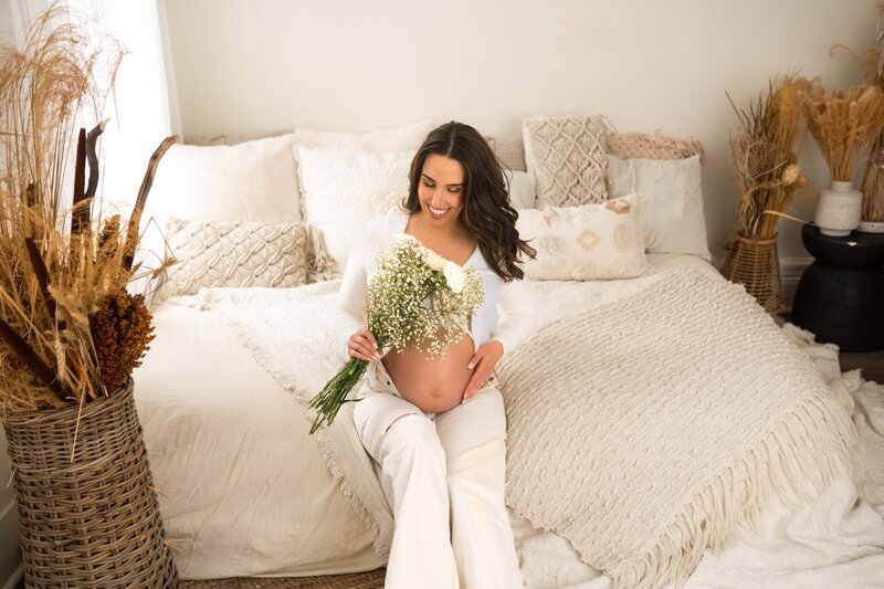 mom sitting on bed holding baby belly and looking at white flowers in her hand