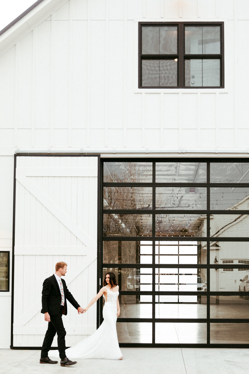 Bride and Groom walking across front of white barn in front of large glass door.
