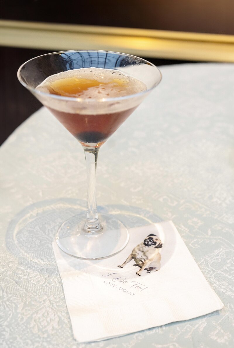 A martini glass filled with a brown cocktail sits on a light-patterned tablecloth. Next to it, a napkin displays a drawing of a small dog and the words Love, Dolly.