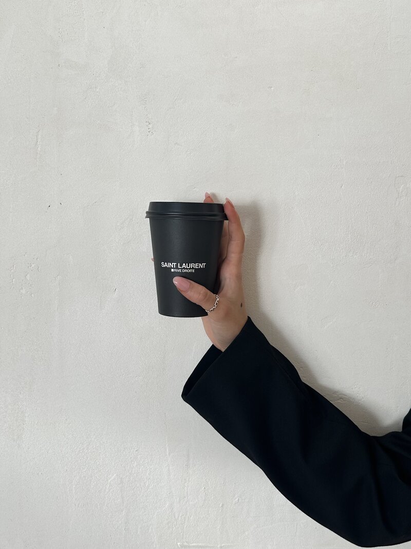 A hand with a minimalist silver ring holds a sleek black coffee cup with the logo "Saint Laurent Rive Droite" against a light, textured wall. The person is wearing a black sleeve, creating a stylish monochrome aesthetic.