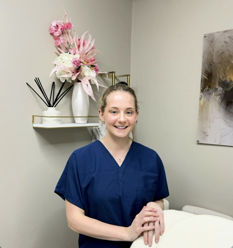 Female clinician in navy scrubs standing and smiling in a treatment room with floral decor on a wall shelf