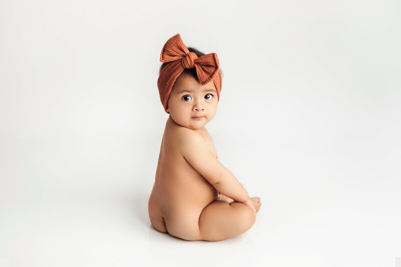 An adorable baby with dark hair sits on a white background, looking over their shoulder with a sweet expression. The baby is naked, except for a large, rust-colored headband with a big bow. The clean, minimalist setting emphasizes the baby's cute and curious demeanor.